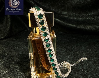 May include: A silver chain bracelet with emerald green rhinestones. The bracelet is laying on top of a glass bottle of perfume.
