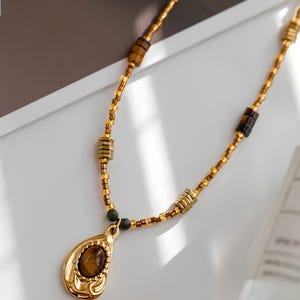 May include: A gold necklace with a teardrop-shaped pendant featuring a brown gemstone. The necklace is made of small gold beads and brown square beads.