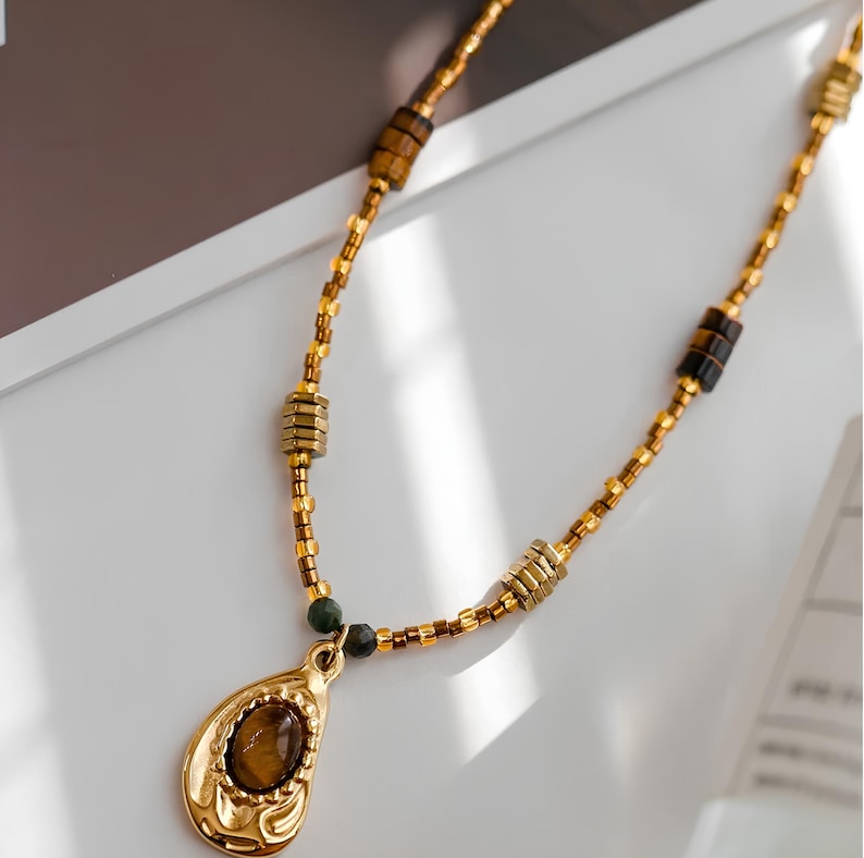 May include: A gold necklace with a teardrop-shaped pendant featuring a brown gemstone. The necklace is made of small gold beads and brown square beads.