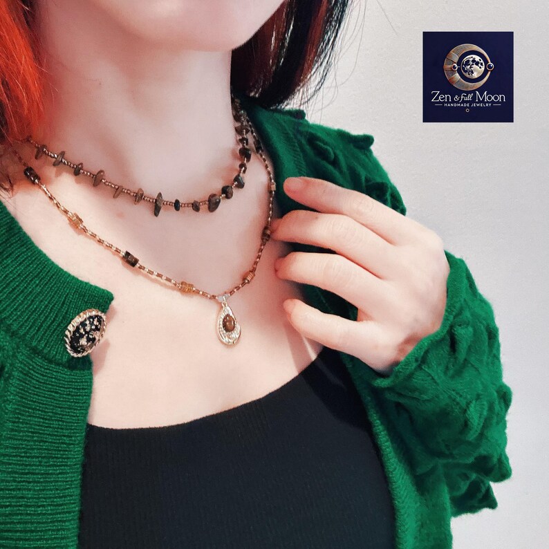May include: A woman wearing a green cardigan and a black tank top is wearing two necklaces. The top necklace is made of brown beads and the bottom necklace is a silver chain with a brown stone pendant. The pendant is shaped like a teardrop. The text "Zen & Full Moon - Handmade Jewelry -" is visible in the top right corner of the image.