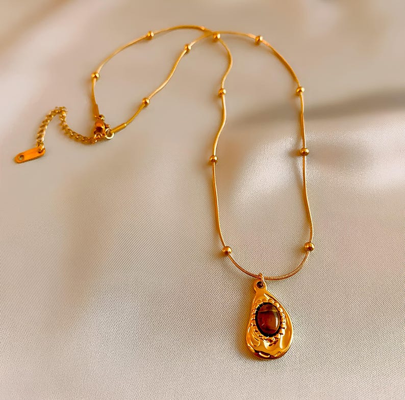 May include: A gold chain necklace with a teardrop-shaped pendant featuring a brown gemstone. The chain has small gold beads.