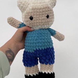 Puede incluir: Muñeca de ganchillo hecha a mano con forma de personaje de dibujos animados. La muñeca tiene una cabeza color crema con ojos negros y orejas pequeñas. Viste una camisa y pantalones cortos azules, con brazos y piernas color beige y zapatos negros. La muñeca es suave y afelpada.