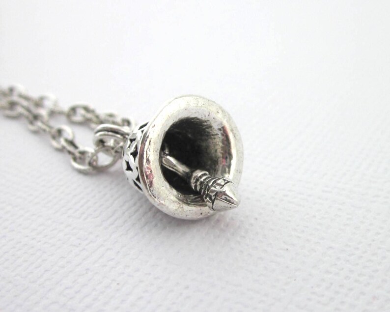 May include: A silver-coloured metal bell pendant attached to a matching link chain. The bell features a detailed, textured exterior surface and a central clapper visible inside the hollow opening. The metal has a polished yet slightly oxidised finish.