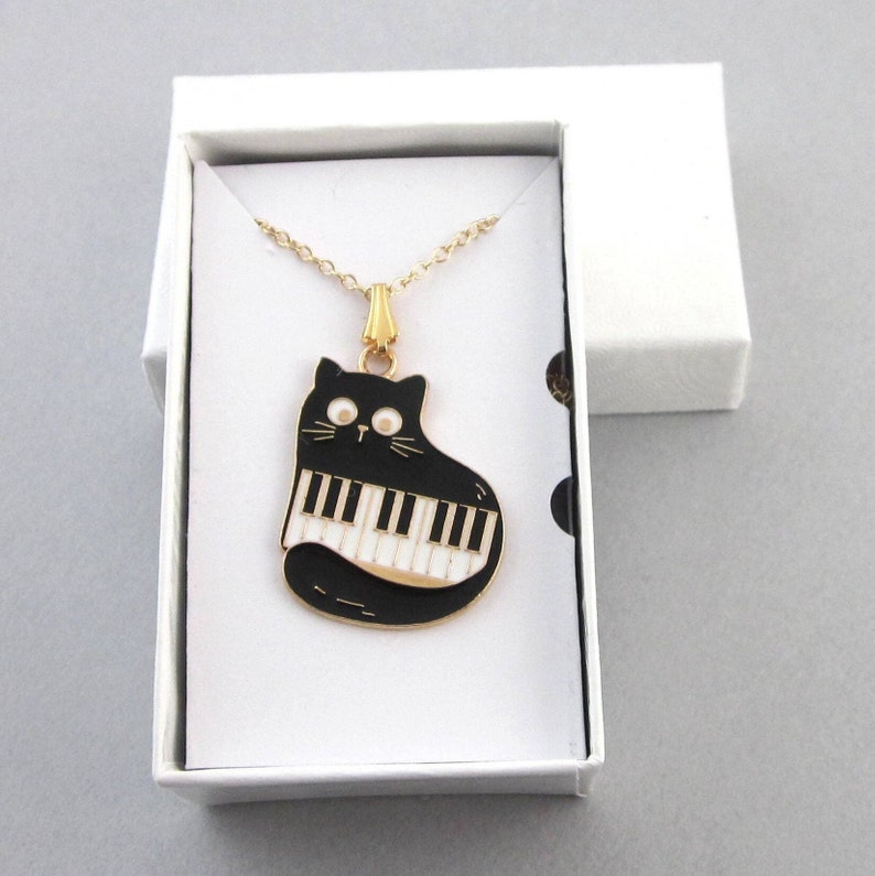 May include: A gold necklace with a black cat charm. The cat is playing a white piano. The charm is in a white box with a gold lid.
