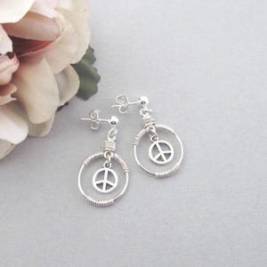 May include: Silver-toned peace sign earrings. Each earring features a circular wire frame with a dangling peace symbol. The earrings have a post-back closure and are displayed on a light gray surface.
