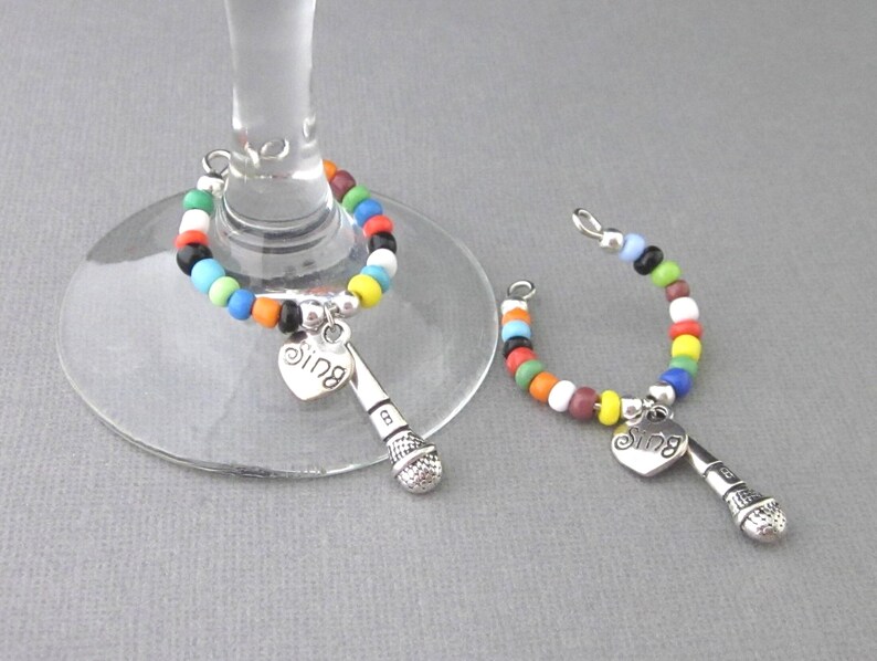 May include: Two colorful beaded wine glass charms with a microphone and heart charm that says "Sing".