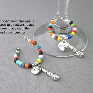 May include: Two colorful beaded wine glass charms with a silver microphone and heart charm with the word "Sing" on it.