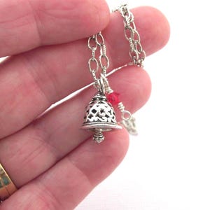 May include: A silver-toned metal bell pendant featuring an intricate filigree cutout pattern, suspended from a matching silver-toned chain. A small, bright red bead is attached to the chain near the top of the bell, adding a pop of colour.