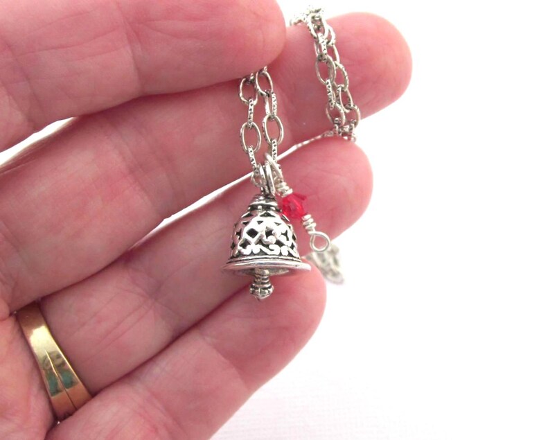 May include: A silver-toned metal bell pendant featuring an intricate filigree cutout pattern, suspended from a matching silver-toned chain. A small, bright red bead is attached to the chain near the top of the bell, adding a pop of colour.
