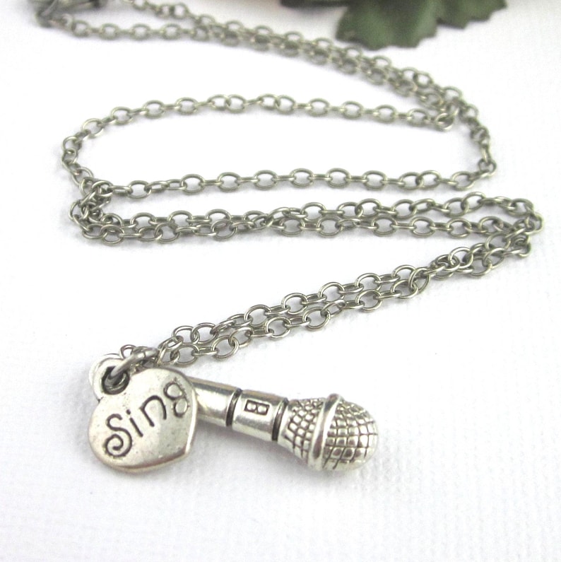 Dimensional microphone charm necklace with separate heart charm showing the engraved word, Sing and a music note on the other side. The mic has realistic features. Charm 27mm x 7mm, heart charm 12mm x 10mm. Chain lengths 18, 17, or 16 inches.