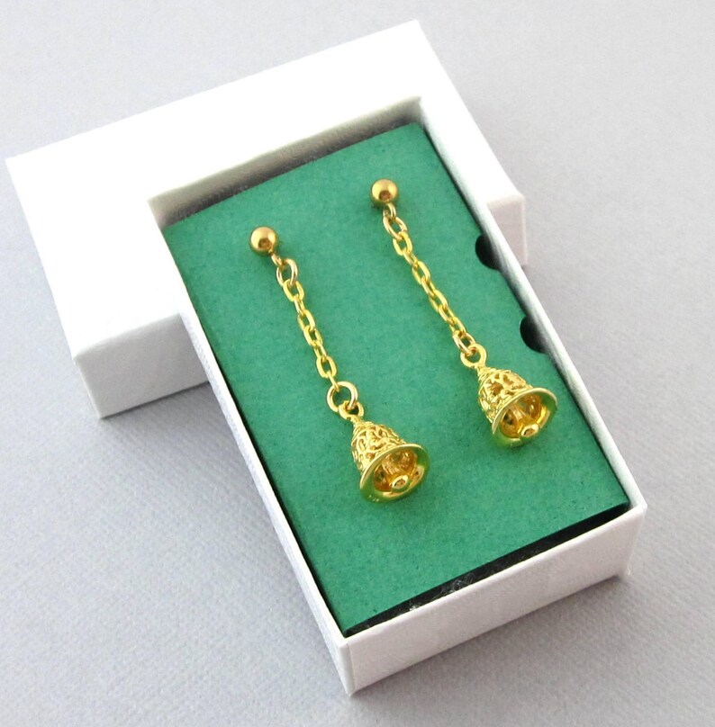 May include: A pair of gold dangle earrings with a bell charm. The earrings are displayed on a green velvet background in a white box.