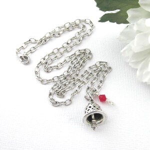 May include: A silver-toned metal chain necklace featuring a small, intricate bell-shaped pendant with cut-out detailing. A single, small, faceted red bead is attached to the jump ring near the bell, adding a subtle pop of colour to the piece.