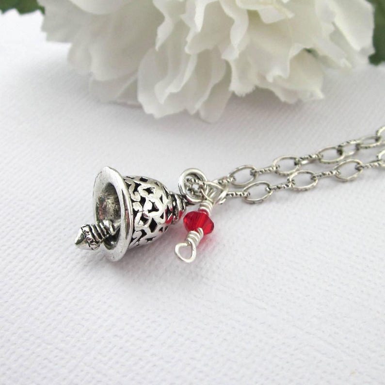 May include: A silver-toned necklace featuring a bell-shaped pendant with an intricate cutout pattern. The pendant is attached to a silver chain and accented with a small, faceted red bead secured by a silver wire wrap detail.