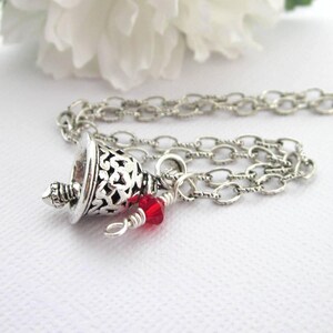 May include: A silver-toned necklace featuring a bell-shaped pendant with an intricate cut-out filigree pattern. A small, faceted, bright red crystal bead is attached to the chain near the pendant with silver wire wrapping.