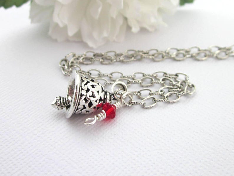 May include: A silver-toned necklace featuring a bell-shaped pendant with an intricate cut-out filigree pattern. A small, faceted, bright red crystal bead is attached to the chain near the pendant with silver wire wrapping.