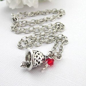 May include: A silver-toned necklace featuring a delicate chain and a bell-shaped pendant with an intricate cut-out pattern. A small, faceted red crystal bead is attached to the jump ring near the top of the bell, adding a touch of colour.