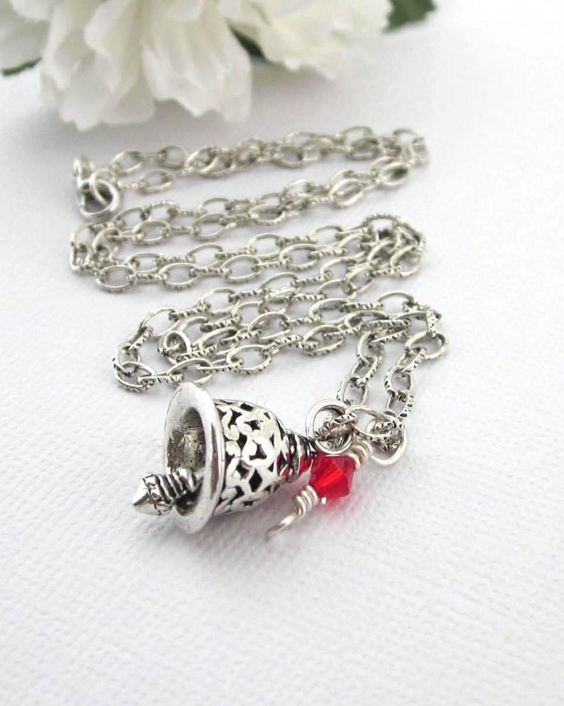 May include: A silver-toned necklace featuring a delicate chain and a bell-shaped pendant with an intricate cut-out pattern. A small, faceted red crystal bead is attached to the jump ring near the top of the bell, adding a touch of colour.