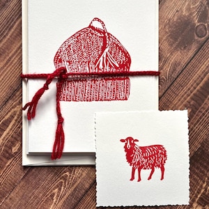 May include: A collection of white cards featuring a red print of a winter hat and a small square card with a red sheep illustration. The cards are bound with red twine, set on a wooden surface.