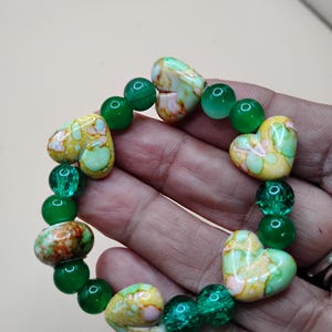 May include: A beaded bracelet featuring heart-shaped and round beads. The heart-shaped beads have a yellow, green, and brown pattern. The round beads are a solid green color. The bracelet is a handmade jewelry item.