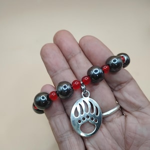Könnte beinhalten: Ein Armband mit dunkelgrauen, kugelförmigen Perlen und kleinen roten Perlen. Ein silberfarbener Anhänger mit einem Bärenpfotenabdruck hängt an dem Armband. Das Armband wird vor einem neutralen Hintergrund gezeigt.