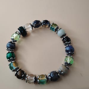 Op de afbeelding: Een armband met kleurrijke kralen met ronde kralen in groen, blauw, bruin en helder. De kralen worden gescheiden door zilverkleurige metalen accenten. Getoond tegen een neutrale achtergrond.