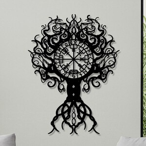 Peut inclure: Décoration murale en métal noir représentant un arbre avec des branches et des racines complexes. Le design intègre une rose des vents au centre, évoquant un thème viking ou nordique. L'œuvre est fixée sur un mur clair, créant un contraste saisissant.