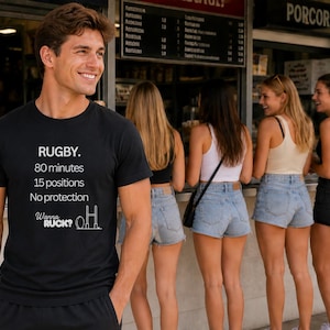 Puede incluir: Camiseta negra con texto blanco: "RUGBY. 80 minutos, 15 posiciones, Sin protección. Wanna RUCK?" Un hombre sonríe frente a un grupo de mujeres. El fondo incluye un puesto de comida y botellas de bebidas.