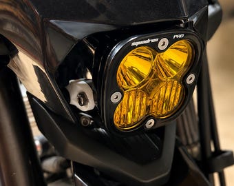 May include: A black motorcycle headlight assembly featuring a square-shaped light with four amber-colored lenses. The housing is marked with the text 'squadron' and 'PRO' in white lettering, secured by four silver-toned circular bolts.