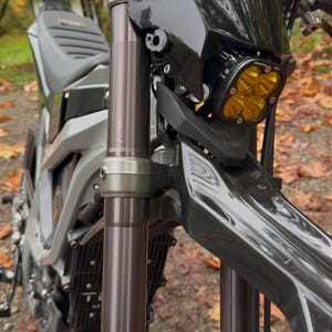 May include: Close-up of a black motorcycle with a black headlight and a yellow LED light. The motorcycle is parked on a gravel path with fallen leaves.