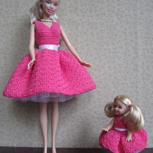 May include: Two Barbie dolls wearing pink crocheted dresses with white sashes and white tulle underskirts. The dolls have blonde hair and blue eyes. The larger doll is standing on a dark surface, while the smaller doll is standing in front of her.