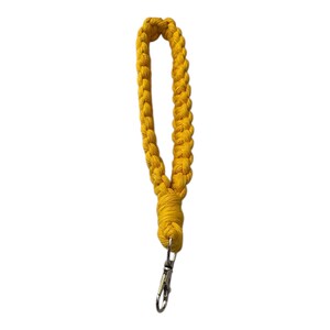 Puede incluir: Una correa de muñeca de macramé amarilla con un cierre de metal plateado. La correa está hecha de un cordón amarillo trenzado. La correa de muñeca está diseñada para sujetarse a un llavero u otros objetos pequeños.