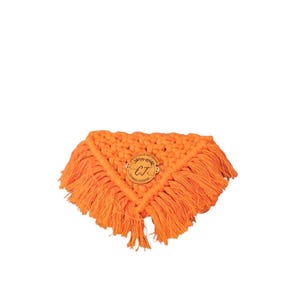Puede incluir: Un clutch de macramé naranja con forma triangular y flecos. Una pequeña etiqueta redonda de madera con las iniciales "CJ" está adherida al centro. El clutch está hecho de material tejido.