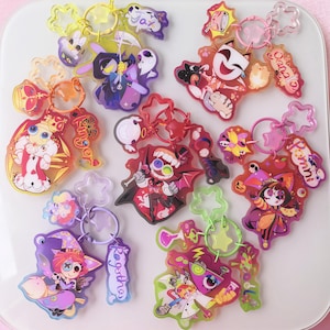 May include: A collection of seven colorful acrylic keychains featuring various cartoon characters, each attached to a star-shaped clasp. The designs include figures with names like Kinger, Jax, Caine, Pomni, Ragatha, and others, rendered in vibrant hues with matching translucent star-shaped rings.