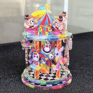 May include: A multi-tiered acrylic carousel display featuring The Amazing Digital Circus branding. The structure includes colourful character charms, a central tent-like top, and a base with a black and white checkered pattern and rainbow accents.