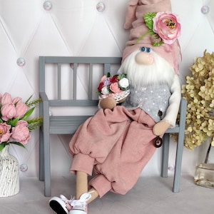 May include: A plush gnome doll with a pink hat and pants, seated on a gray bench. The gnome has a white beard, blue eyes, and a pink nose. It holds a basket of flowers and wears white sneakers with pink accents.