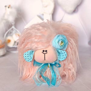 May include: A plush toy sheep with long, shaggy light pink fur. The sheep has a beige face with black embroidered eyes and nose, turquoise polka-dot ears, a turquoise fabric flower on its head, and a turquoise ribbon tied around its neck.
