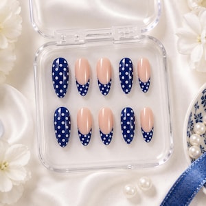 May include: A set of ten false nails in a clear plastic case. The nails feature a navy blue base with white polka dots and a nude-coloured upper section. The nails are oval-shaped.