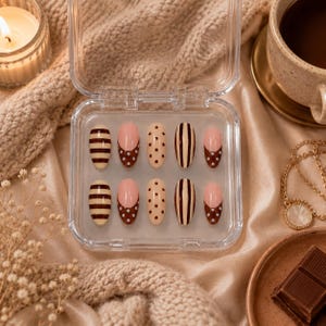 May include: A set of ten press-on nails in a clear plastic case, featuring a mix of designs including horizontal brown stripes, brown French tips with white dots, and cream-coloured nails with brown polka dots or vertical brown stripes.
