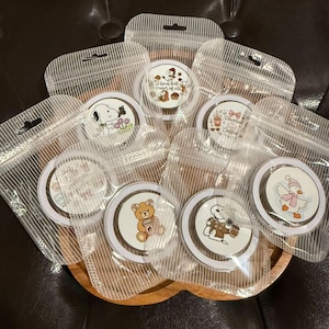 May include: A collection of circular stickers packaged in individual clear plastic pouches. The stickers feature various cartoon illustrations including a dog with flowers, a teddy bear holding a cup, a duck in a winter hat, and text designs.