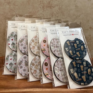 May include: Six sets of two circular car coasters packaged in clear plastic sleeves with white header cards labeled Car Coasters Handmade with love. Each set features distinct patterns including coffee cups, flowers, ghosts, and donuts.