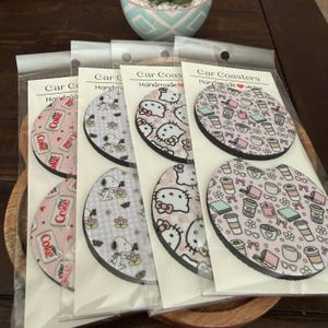 May include: A collection of handmade car coasters in clear plastic packaging. The coasters feature various designs, including Coca-Cola logos, Snoopy, Hello Kitty, and coffee-themed patterns. The coasters are displayed on a wooden surface.