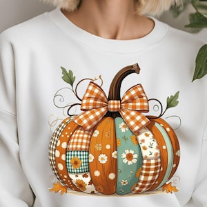 以下が含まれることがあります： カラフルなカボチャのデザインが施された白いスウェットシャツ。カボチャは、水玉模様、ギンガムチェック、花柄など、さまざまなパターンで装飾されています。オレンジと白の大きなチェック柄のリボンが上にあり、緑の葉とつるが周りにあります。