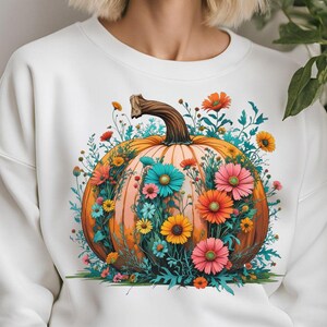 May include: White sweatshirt featuring a large, detailed illustration of a pumpkin adorned with colorful flowers. The pumpkin is orange with a brown stem, and the flowers are various colors, including pink, orange, and teal. The design is centered on the front.