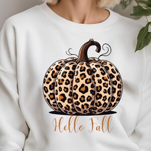 Peut inclure: Sweat-shirt blanc avec un motif de citrouille. La citrouille a une tige marron et un motif léopard marron et beige. Les mots "Hello Fall" sont écrits en cursive en dessous. Un article sur le thème de l'automne.