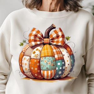 May include: Cream-colored sweatshirt featuring a colorful patchwork pumpkin design. The pumpkin has a large plaid bow and is composed of various patterned fabric pieces in orange, yellow, blue, and white. A fall-themed graphic.
