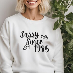 May include: White sweatshirt with black text that reads "Sassy Since 1985" with two small hearts above the year.