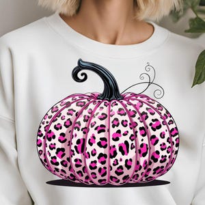 May include: A white sweatshirt with a pink leopard print pumpkin design. The pumpkin has a black stem and is decorated with pink and black spots. The design is a festive and stylish autumn or Halloween theme.