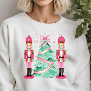 Puede incluir: Sudadera blanca con un árbol de Navidad rosa y verde y dos figuras de cascanueces rosas y rojas.