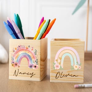 May include: Two wooden pen holders with rainbow designs and names. One is filled with pens in various colours. The rainbow designs feature pastel colours and floral accents. The names "Nancy" and "Olivia" are written in cursive.