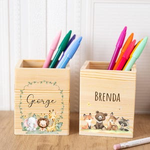 Puede incluir: Dos portalápices de madera con bolígrafos de colores. Un soporte tiene el nombre "George" con una ilustración de un elefante, un león y una jirafa. El otro soporte tiene el nombre "Brenda" con ilustraciones de un zorro, un oso, un ciervo y un mapache.
